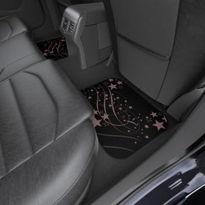 May include: A set of black car floor mats featuring a repeating pattern of light pink stars and swirling lines. The mats are positioned on the floor of a vehicle, covering the footwell area in front of the rear seats and the centre console.