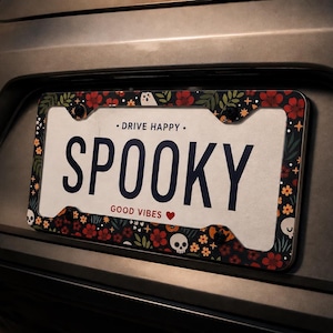 May include: A rectangular license plate frame featuring a black background with a pattern of red flowers, green leaves, small white skulls, and tiny yellow stars. The text DRIVE HAPPY is printed at the top and GOOD VIBES with a red heart at the bottom.