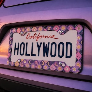 Może przedstawiać: Tablica rejestracyjna stanu Kalifornia z napisem "HOLLYWOOD" w granatowym kolorze. Ramka tablicy ma powtarzający się wz&oacute;r diament&oacute;w, gwiazd i księżyc&oacute;w w kolorze r&oacute;żowym, fioletowym i ż&oacute;łtym. Słowo "California" jest napisane czerwoną czcionką.