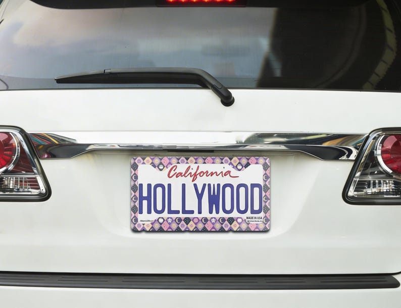 Może przedstawiać: Ozdobna tablica rejestracyjna z napisem "HOLLYWOOD" w dużych, niebieskich literach. Tablica jest umieszczona na białym samochodzie i ma wzorzystą obw&oacute;dkę z r&oacute;żowymi, fioletowymi i białymi geometrycznymi kształtami. Słowo "California" jest napisane kursywą.