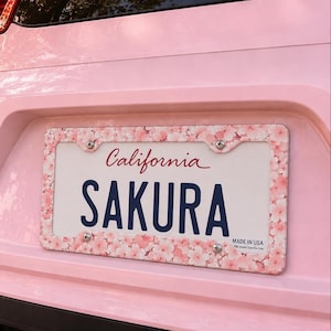 Puede incluir: Un marco de matrícula rectangular con un patrón repetitivo de flores de cerezo de color rosa claro. El marco está montado en un vehículo de color rosa claro y rodea una placa blanca con el texto California y SAKURA en azul oscuro.