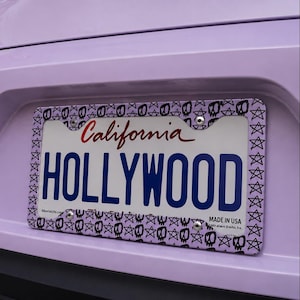 Puede incluir: Un marco de matrícula con un patrón repetido de calaveras y estrellas rodea una placa de matrícula de California. La matrícula dice "HOLLYWOOD" en letras azules. La palabra "California" está en escritura roja.