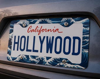 May include: A license plate frame with a white license plate that reads "HOLLYWOOD" in blue block letters. The word "California" is written in red script above the license plate. The frame has a blue and white wave pattern.