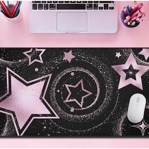 May include: Black rectangular mouse mat featuring a pattern of pink stars and a white mouse. The design includes large and small stars, with a glittery effect. The mouse mat is on a pink surface.