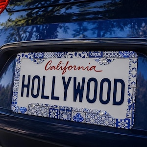 Puede incluir: Matrícula de coche con un marco decorativo con estampado de azulejos azules y blancos. La placa muestra "California" en escritura roja y "HOLLYWOOD" en mayúsculas azul marino.