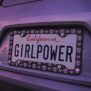 Puede incluir: Una placa de matrícula de California con la palabra "GIRLPOWER" en negro. La placa está enmarcada con un borde negro adornado con calaveras y corazones rosas. El fondo es un coche morado.