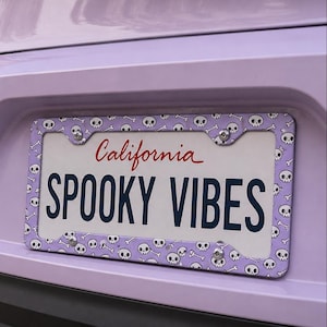 Puede incluir: Un marco de matrícula lila claro con un patrón de calaveras y huesos blancos. La placa muestra "California" en escritura roja y las palabras "SPOOKY VIBES" en azul marino. Un accesorio de coche con temática de Halloween.