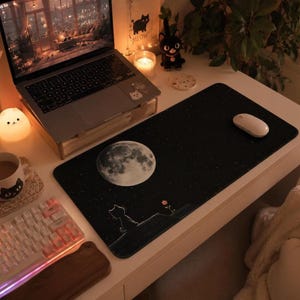 May include: A rectangular black desk mat featuring a large, detailed white full moon in the upper left corner, a small white outline of a cat sitting on a hill, and a tiny orange flower, all set against a starry night sky background.