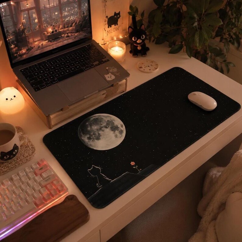 May include: A rectangular black desk mat featuring a large, detailed white full moon in the upper left corner, a small white outline of a cat sitting on a hill, and a tiny orange flower, all set against a starry night sky background.