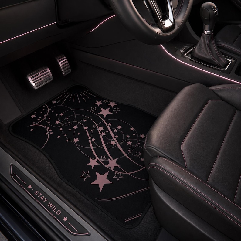 May include: A black car floor mat featuring a light pink graphic design of swirling lines and various sized stars. The mat is positioned in the driver side footwell of a vehicle, which also includes a door sill plate with the text STAY WILD.