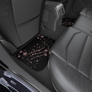 May include: A set of black car floor mats featuring a repeating pattern of light pink stars and swirling lines. The mats are installed in the rear footwell of a vehicle, positioned between the front seat back and the rear leather bench seat.