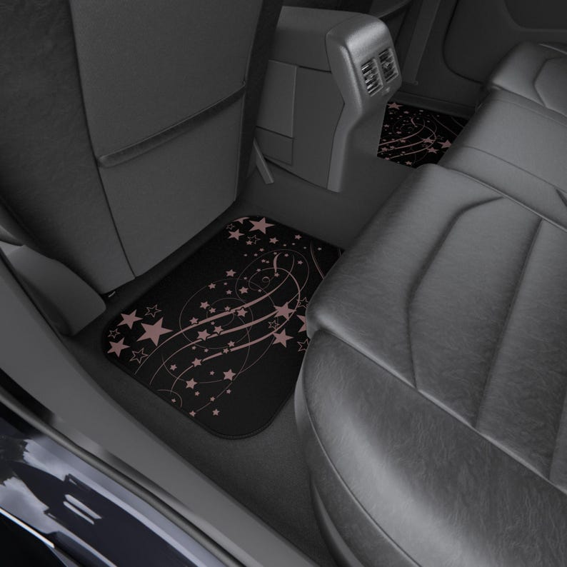May include: A set of black car floor mats featuring a repeating pattern of light pink stars and swirling lines. The mats are installed in the rear footwell of a vehicle, positioned between the front seat back and the rear leather bench seat.