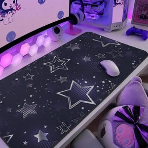 Puede incluir: Alfombrilla de escritorio negra con un patrón de estrellas plateadas y un ratón de ordenador blanco. La alfombrilla es rectangular y presenta estrellas plateadas de varios tamaños. Un personaje de peluche morado es visible.