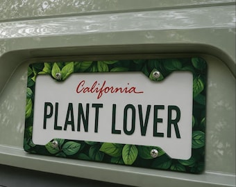 May include: A California license plate frame featuring a green leaf pattern. The plate displays "PLANT LOVER" in bold, black text, with "California" in red script. The frame is attached to a vehicle, likely a car or truck.