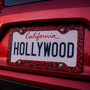 Puede incluir: Un marco rectangular para matrícula con un patrón repetitivo de estrellas negras y rojas. El marco está sujeto con cuatro tornillos plateados alrededor de una matrícula blanca de California con la palabra HOLLYWOOD en letras azul oscuro.