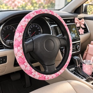 Puede incluir: Una funda para volante con un patrón repetitivo de flores de cerezo en tonos rosa y rosa claro. La funda está instalada en un volante negro dentro de un vehículo, junto a un accesorio de lazo rosa y un vaso rosa a juego.