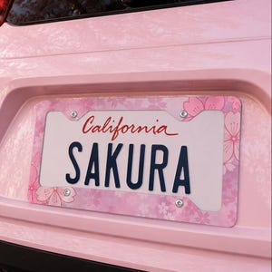 Puede incluir: Un marco rosa para matrícula con un diseño floral y la palabra "SAKURA" en azul marino. La matrícula dice "California" en escritura roja. El marco está montado en un coche rosa.
