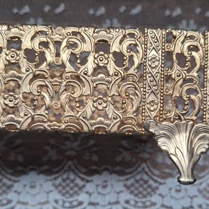 May include: A rectangular gold-toned metal tray featuring an intricate openwork filigree pattern of swirling scrolls and small floral motifs. The tray is supported by a decorative, ornate metal foot extending from the side corner.