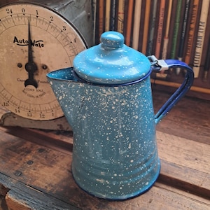 May include: A blue enamel coffee pot with a white speckled pattern, a matching lid with a small knob, a curved handle, and a wide spout. The pot features a dark blue rim and a tapered cylindrical body, showcasing a classic kitchenware design.
