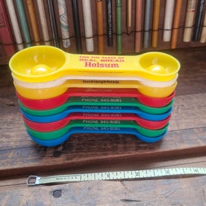 May include: A stack of six double-ended plastic measuring spoons in yellow, white, red, green, and blue. The top yellow spoon features the text FOR THE TASTE OF REAL BREAD Holsum. A white measuring tape shows a length of 4 inches.