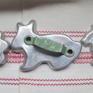 May include: Three metal cookie cutters including a scalloped circular shape with a red handle, a larger dog-shaped cutter with a green metal handle, and a smaller dog-shaped cutter with a red handle, all featuring silver-toned metal construction.