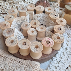 May include: A collection of light-coloured wooden spools arranged on a circular wooden base, interspersed with white lace trim. Each spool features a printed paper label on top with text including Belding Corticelli, shade numbers, and prices.