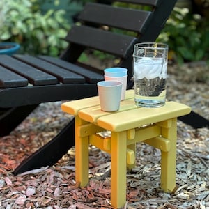 May include: A small yellow side table with a slatted top surface. On the table sits a clear glass pitcher filled with water and ice cubes, alongside two small, light-colored cups, one stacked inside the other, positioned next to the pitcher.