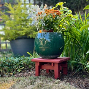May include: A dark green, round ceramic planter filled with orange and white flowers sits on a small, red wooden plant stand. The scene is set outdoors with lush greenery in the background.