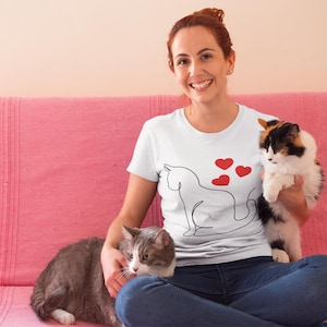 May include: A woman wearing a white t-shirt with a line drawing of a cat and hearts on it. She is sitting on a pink couch with two cats. One cat is sitting on her lap and the other is sitting on the couch.