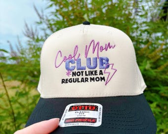 Könnte beinhalten: Beige und schwarze Baseballkappe mit dem Text "Cool Mom Club - Not Like A Regular Mom" in Lila und Blau. Die Kappe hat einen schwarzen Schirm und einen schwarzen Knopf oben. Der Hut ist ein Mid-Profile, OSFM (One Size Fits Most) Hut.
