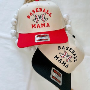 May include: Two baseball caps with the text "Baseball Mama" and a red bow with crossed baseball bats embroidered on the front. One cap has a red brim and the other has a black brim.