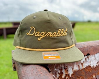 May include: Olive green baseball cap with the embroidered word "Dagnabbit" in gold thread. The cap has a flat brim and a gold rope detail along the brim. A small orange and white tag is visible on the brim.