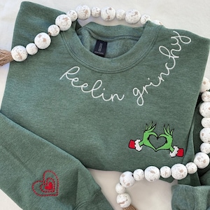 Puede incluir: Sudadera verde con la frase "Feelin Grinchy" bordada en blanco. Presenta un diseño de corazón de manos del Grinch y un corazón rojo en la manga. Una guirnalda de cuentas blancas y marrones decora la parte superior.