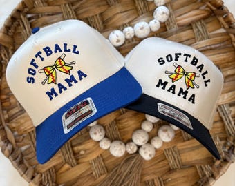 May include: Two beige baseball caps with black brims. The caps have an embroidered design of a yellow softball with a red bow and crossed bats. The text "Softball Mama" is embroidered in blue on the front of each cap. One cap has a blue brim and the other has a black brim.