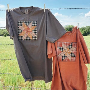 Pode incluir: Duas camisetas penduradas em um varal. Uma é cinza escuro e a outra laranja queimado. Cada camisa apresenta um design de patchwork em forma de estrela no centro, com uma mistura de padrões e cores. O fundo mostra um campo verde e um céu azul.