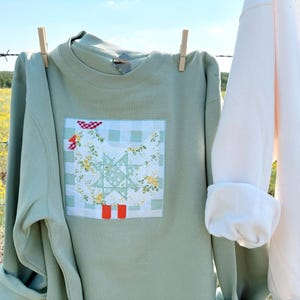 Könnte beinhalten: Hellgrünes Sweatshirt mit einem Patchwork-Design einer Henne mit roten Stiefeln und floralen Mustern. Das Sweatshirt hängt an einer Wäscheleine mit einer Wäscheklammer. Ein weiteres weißes Sweatshirt ist sichtbar.