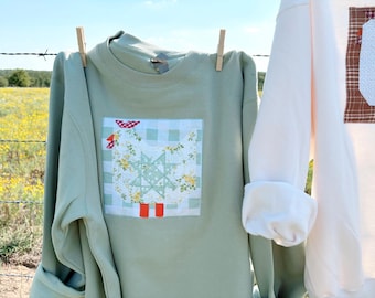 Könnte beinhalten: Hellgrünes Sweatshirt mit einem Patchwork-Design einer Henne mit roten Stiefeln und floralen Mustern. Das Sweatshirt hängt an einer Wäscheleine mit einer Wäscheklammer. Ein weiteres weißes Sweatshirt ist sichtbar.