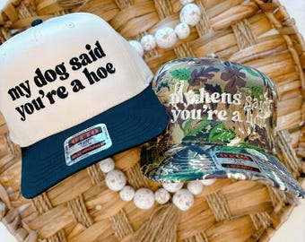 May include: Two trucker hats with funny sayings. The first hat is beige with black lettering that says "my dog said you're a hoe". The second hat is camouflage with black lettering that says "my hens said you're a hoe".