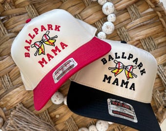 May include: Two baseball caps with a beige crown and black or red brim. Both caps feature an embroidered design of a baseball bat and bow with the text "Ballpark Mama".