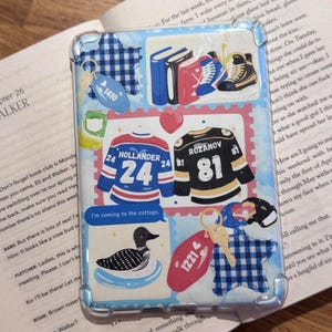 May include: A clear phone case with a light blue gingham pattern and various stickers. Stickers include hockey jerseys, books, a loon, and keychains. The text "I'm coming to the cottage" is also visible.
