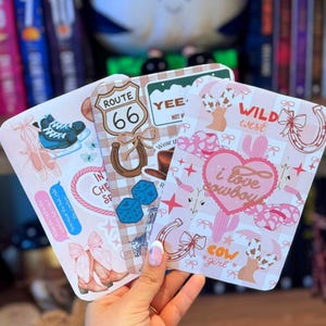 May include: Three decorative cards with a Western theme. One card features a Route 66 sign and dice, another has a cowboy hat and the text "I love cowboys", and the third has a pair of cowboy boots and ice skates. The cards are pink and white.