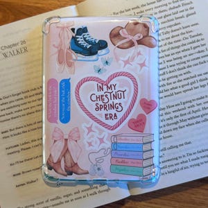 May include: A clear phone case with a pink background featuring illustrations of ballet shoes, ice skates, a cowboy hat, butterflies, and cowboy boots. The case has text that reads "In My Chestnut Springs Era."