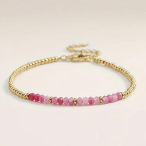 May include: A gold bracelet with a beaded chain and a row of pink and fuchsia gemstones. The bracelet has a lobster clasp. The gemstones are arranged in a gradient of shades. The bracelet is suitable for everyday wear.