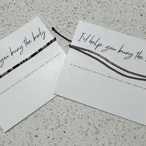 May include: Two friendship bracelets on white cards. One bracelet is black with gold beads, the other is purple with gold beads. The cards read "I'd help you bury the body" in black script.