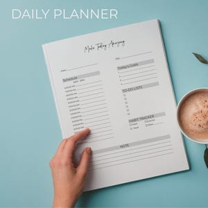 Può includere: Un'agenda giornaliera bianca con il testo "DAILY PLANNER" e "Make Today Amazing". L'agenda include sezioni per la programmazione, gli obiettivi, le liste di cose da fare e un tracker delle abitudini. Una tazza di caffè e un rametto di foglie sono su una superficie azzurra.