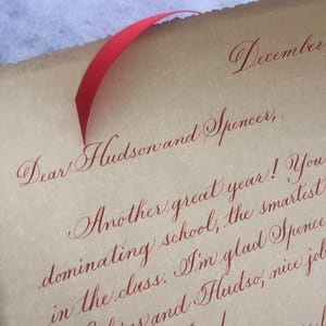 May include: A handwritten letter on parchment paper with red ribbon tied around it. The letter reads "December, Dear Hudson and Spencer, Another great year! You dominating school, the smartest in the class. I'm glad Spence Hoskins and Hudso, nice job Spelling Bee! I believe you are..."