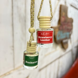 May include: Two car air fresheners hanging from a gold and beige string. The air fresheners are in glass bottles with wooden tops. The top air freshener is labeled "rustic charm candles Strawberry Leather" and the bottom air freshener is labeled "rustic charm candles Buckaroo".