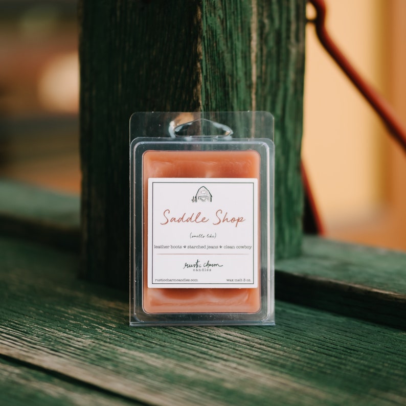 May include: A small, clear plastic container with a brown wax melt inside. The wax melt is labeled "Saddle Shop" with the text "(smells like)" and "leather boots starched jeans clean cowboy". The label also includes the text "rust charm candles" and "wax melt 3 oz."