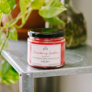 May include: A red glass candle jar with a black lid. The label reads "Strawberry Leather (smells like) fresh strawberry + leather + picnic by the river" and "rustic chum candles" with a website address. The candle is a soy blend and is 12 oz.