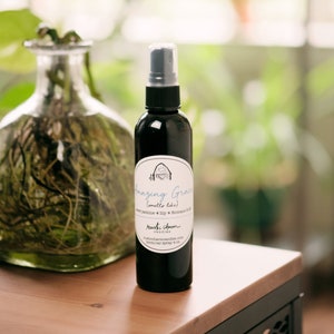 May include: A black glass bottle with a spray nozzle and a white label that reads "Amazing Grace (smells like) Sweet Jasmine + Lily + Roman 6:23 Rustic Charm Candles rusticcharmcandles.com room/car spray 4 oz"